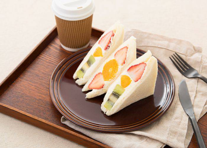 Three fruit sandwich halves on a plate, surrounded by cutlery, napkins and a to-go coffee.